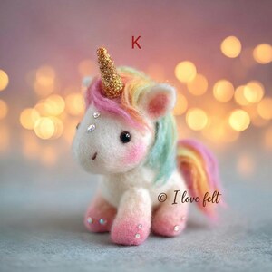 Op de afbeelding: Een handgemaakte vilten eenhoorn met een gouden hoorn, regenboogmanen en -staart en roze accenten. De eenhoorn heeft een wit lichaam, zwarte ogen en is versierd met kleine strass-steentjes. De letter "K" is zichtbaar op de achtergrond, evenals het copyright en de tekst "I love felt".