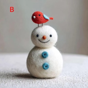 Puede incluir: Una figura de muñeco de nieve de lana fieltrada blanca con un pájaro rojo posado en la cabeza. El muñeco tiene una pequeña nariz de zanahoria naranja, dos ojos negros, una sonrisa negra cosida y dos botones azules. La letra B se ve en rojo.