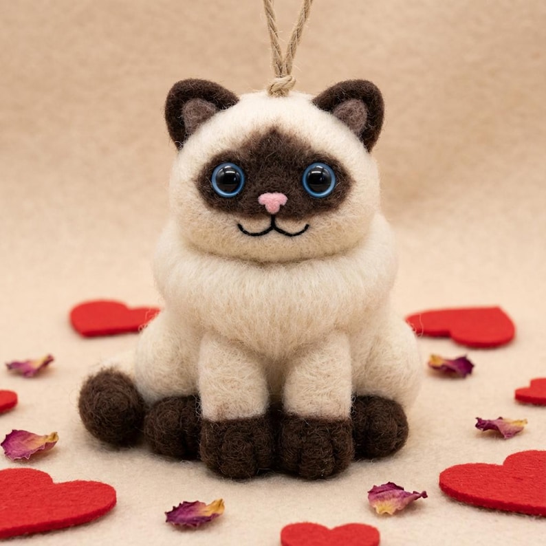 May include: A handmade felt Siamese cat ornament with a cream-colored body, brown markings, and blue eyes. The cat is sitting with its legs forward and has a string for hanging. Red felt hearts and rose petals are scattered around the cat.