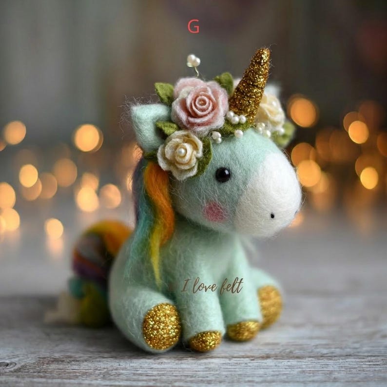Op de afbeelding: Een vilten eenhoornbeeldje in pastelgroen met een gouden glitterhoorn en hoeven. Het heeft een regenboogmanen en -staart en een bloemenkrans met roze en witte rozen. De tekst "I love felt" staat op de zijkant.
