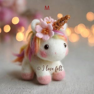 Op de afbeelding: Handgemaakte eenhoornfiguur van vilt met een wit lichaam, roze hoeven en een regenboogmanen. Het heeft een gouden glitterhoorn en een bloemenkrans. De tekst "&copy; I love felt" is zichtbaar op het lichaam van de eenhoorn.