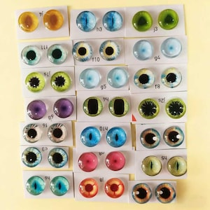 May include: A collection of glass eye cabochons in various colors and designs. The eyes feature a range of colors, including blue, green, brown, and purple, with detailed iris and pupil patterns. Each pair is mounted on a small white card.