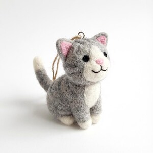 May include: A gray and white felt cat ornament with pink inner ears and a pink nose. The cat has black eyes and a smiling mouth. A loop of twine is attached for hanging. The cat is a decorative item.