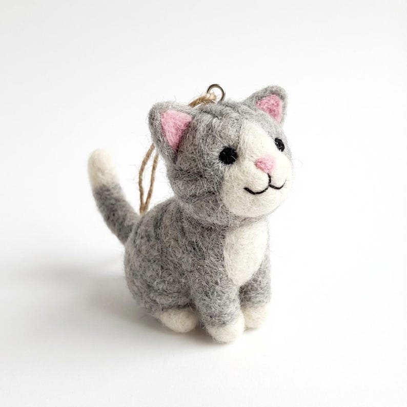 May include: A gray and white felt cat ornament with pink inner ears and a pink nose. The cat has black eyes and a smiling mouth. A loop of twine is attached for hanging. The cat is a decorative item.