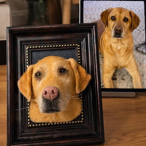 May include: A framed, felted sculpture of a yellow Labrador Retriever dog. The dog's head and face are the focal point, with a pink nose and detailed features. The frame is dark brown with gold-colored studs. A tablet displays a photo of a similar dog.