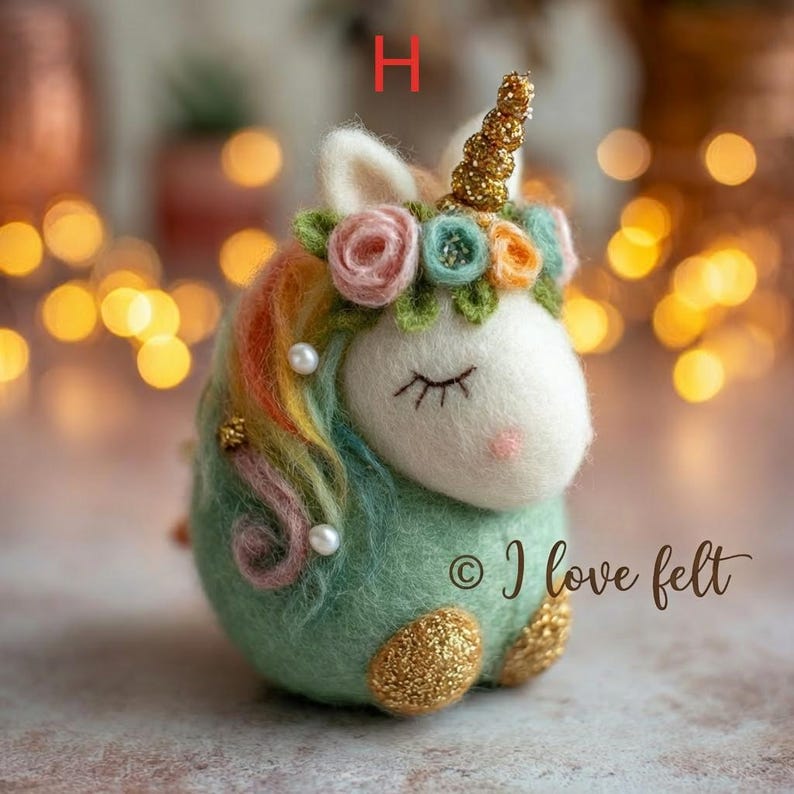 Op de afbeelding: Een vilten eenhoornfiguur met een wit gezicht, gesloten ogen en een roze neus. Het heeft een gouden hoorn, een bloemenkrans en een regenboogmanen. De body is groen met gouden glitteraccenten. De tekst "&copy; I love felt" is zichtbaar.