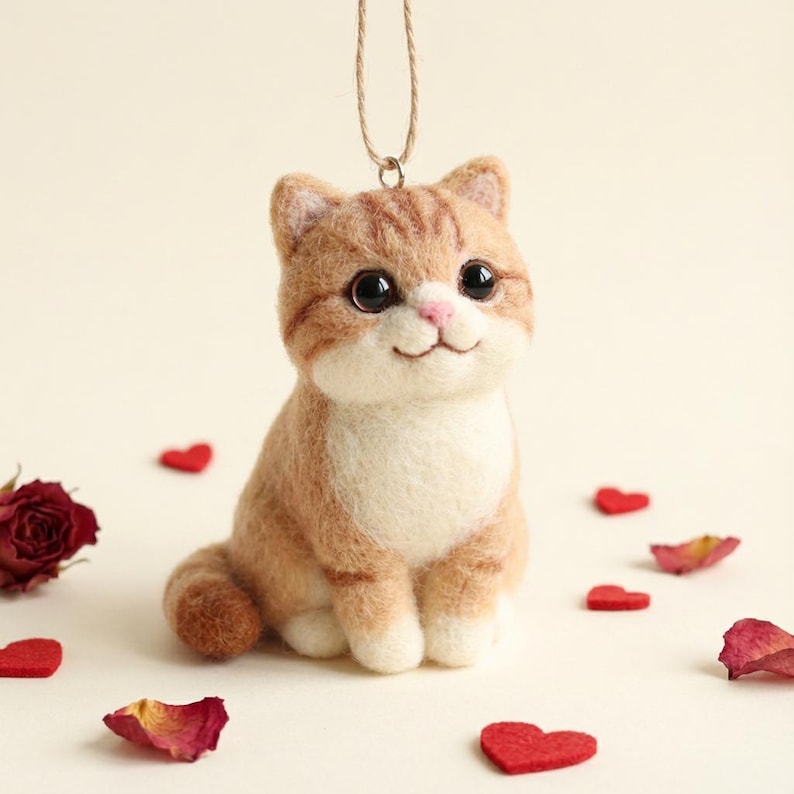 May include: A handmade felt cat ornament in shades of orange and white, with large black eyes and a pink nose. The cat is sitting, with a string for hanging. Red heart-shaped decorations and rose petals are scattered around the cat.