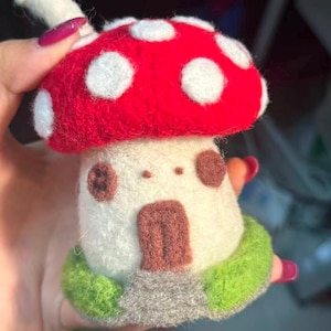 May include: A needle-felted mushroom house figurine featuring a bright red cap with white polka dots, a cream-colored stem with two brown circular windows, a brown arched door, and a green grassy base with a small gray stone path at the front.