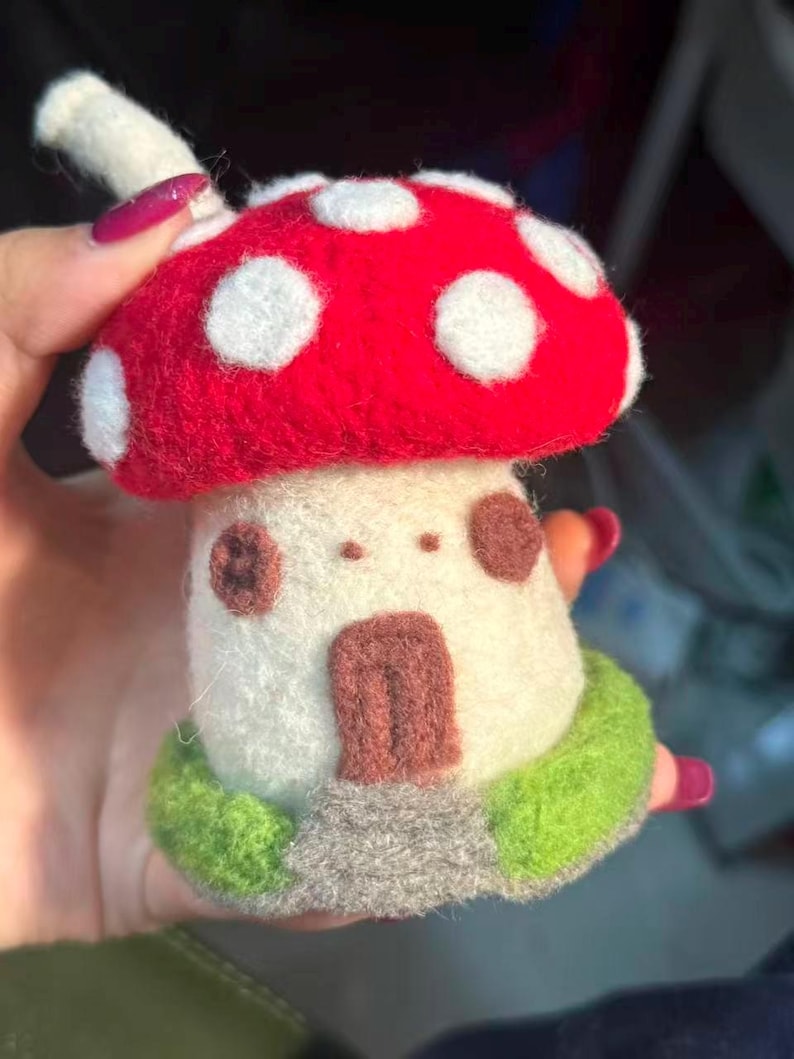 May include: A needle-felted mushroom house figurine featuring a bright red cap with white polka dots, a cream-colored stem with two brown circular windows, a brown arched door, and a green grassy base with a small gray stone path at the front.