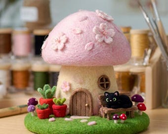 May include: A needle-felted mushroom house with a light pink cap adorned with cherry blossoms, a cream-colored stem, a small wooden door, and a circular window. A black cat sits on a bench next to red potted succulents and small colorful mushrooms.