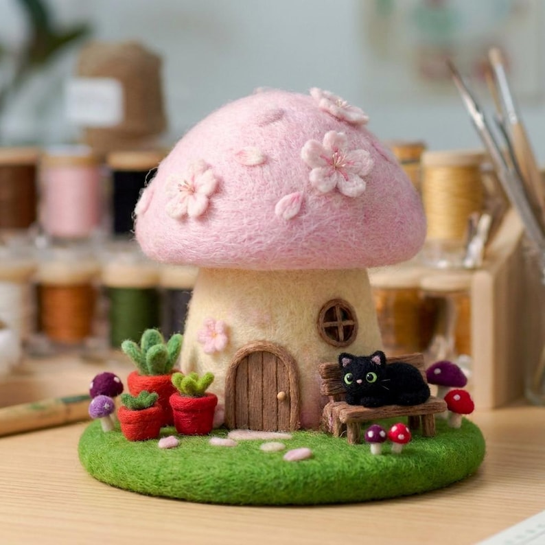 May include: A needle-felted mushroom house with a light pink cap adorned with cherry blossoms, a cream-colored stem, a small wooden door, and a circular window. A black cat sits on a bench next to red potted succulents and small colorful mushrooms.