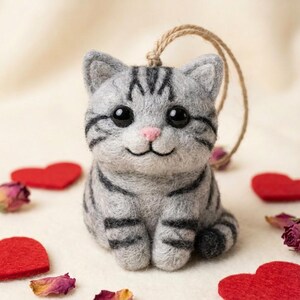 May include: A gray and black striped felt cat ornament with a jute string for hanging. The cat has large black eyes, a pink nose, and a smiling mouth. Red felt hearts and dried rose petals are scattered around the cat.