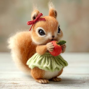 Può includere: Una figura di scoiattolo fatta a mano in feltro, con pelliccia arancione, un fiocco rosso e una gonna verde, che tiene in mano una mela rossa. Lo scoiattolo ha grandi occhi espressivi ed è posizionato su una superficie chiara.