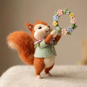 May include: A handcrafted felt squirrel figurine with a fluffy orange tail and a light green jacket. The squirrel holds a floral wreath with pink, blue, and yellow flowers. The figurine has a cheerful expression and is set against a soft background.