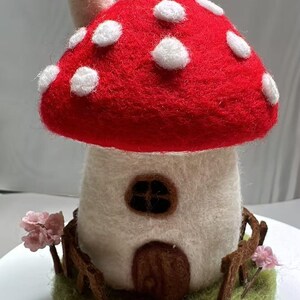 May include: A needle-felted mushroom house sculpture featuring a bright red cap with white spots, a white stem with a brown door and window, a chimney, a brown fence, a green base, and small pink flowers on either side of the structure.