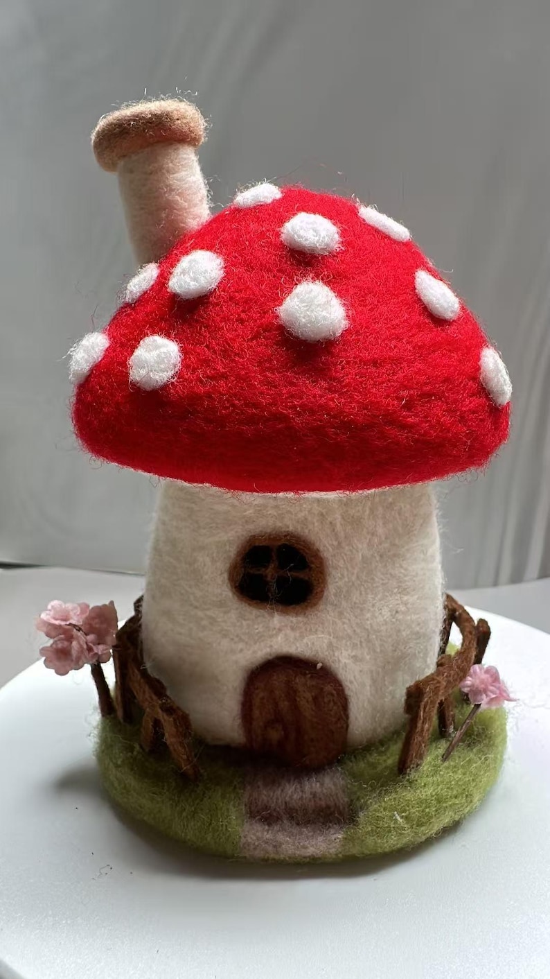May include: A needle-felted mushroom house sculpture featuring a bright red cap with white spots, a white stem with a brown door and window, a chimney, a brown fence, a green base, and small pink flowers on either side of the structure.