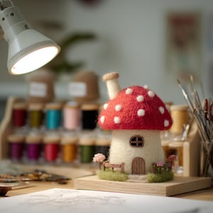 May include: A needle-felted mushroom house sculpture featuring a bright red cap with white polka dots, a cream-colored stem, a small brown arched door, a window, and a tiny fence. The structure sits on a green base with miniature pink flowers.
