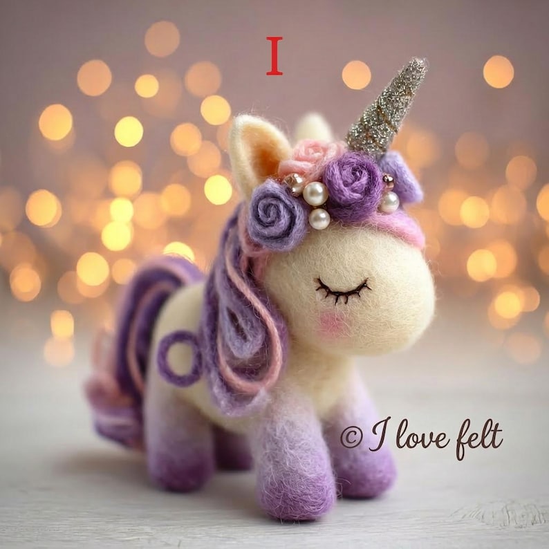 Op de afbeelding: Handgemaakte vilten eenhoornfiguur met een zilveren glitterhoorn, paarse en roze manen en staart, en een bloemenkrans met parels. De eenhoorn heeft gesloten ogen en roze wangen. De tekst "I love felt" is zichtbaar.
