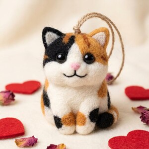 May include: A handmade felt cat figurine with black, orange, and white patches. The cat has a pink nose, black eyes, and a smiling mouth. A twine loop is attached for hanging. Red felt hearts and dried rose petals are scattered around the cat.