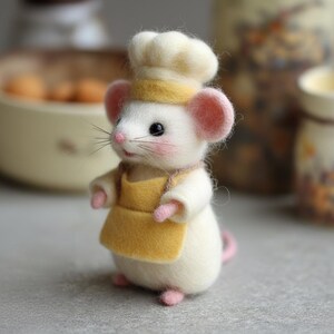 Pu&ograve; includere: Figurina di topo in feltro fatta a mano vestita da chef. Il topo indossa un cappello da chef bianco con una fascia gialla, un grembiule giallo e ha orecchie, naso e piedi rosa. La figura &egrave; su uno sfondo sfocato.