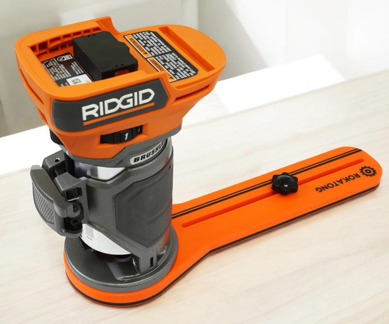 K&ouml;nnte beinhalten: Ein orangefarbener und grauer b&uuml;rstenloser RIDGID-Router mit einem schwarzen Batteriefach. Der Router ist auf einer orangefarbenen Basis mit dem Text "ROKATONG" montiert. Das Werkzeug ist f&uuml;r die Holzbearbeitung und Zimmerei konzipiert.