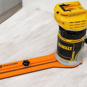 May include: A yellow and black DEWALT brushless power tool with an orange base. The base has the text "ROKATONG" and a black knob. The tool is on a light wood-grain surface.
