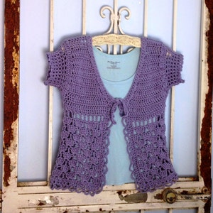 May include: A lavender crocheted cardigan with short sleeves and a tie front. The cardigan is hanging on a white metal hanger.