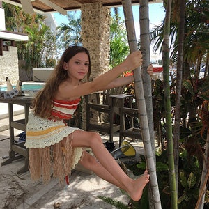 May include: A young girl wearing a crocheted dress with red, yellow, and white stripes and a fringe hem. She is holding onto a tall, thin tree trunk with her right hand. The background is a tropical setting with palm trees and a wooden pergola.