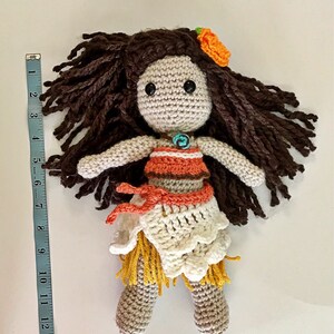 May include: Crocheted doll with brown hair, a flower in her hair, and a white and orange dress. The doll is wearing a turquoise necklace and has brown eyes.