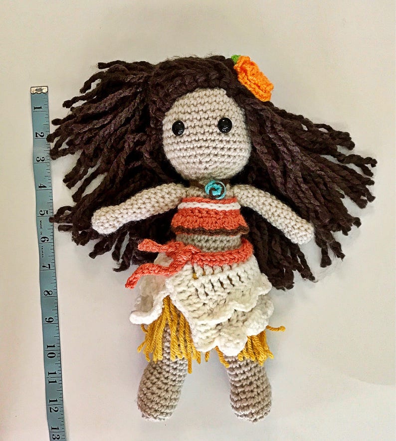 May include: Crocheted doll with brown hair, a flower in her hair, and a white and orange dress. The doll is wearing a turquoise necklace and has brown eyes.