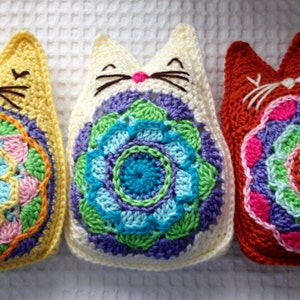Puede incluir: Tres juguetes de crochet con forma de gato con diseños de mandala coloridos. Los gatos son amarillos, blancos y naranjas.