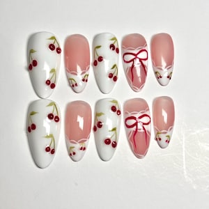 May include: A set of ten press-on nails featuring an almond shape. Some nails have a solid white base with red cherry illustrations and green stems. Other nails feature a translucent pink base with white lace trim and red ribbon bow designs.