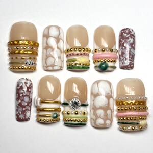 Op de afbeelding: Een set van tien kunstnagels met diverse ontwerpen, waaronder nude bases met gouden metalen bandjes, wit en bruin gespikkelde patronen, kleine parelaccenten, roze en groene horizontale strepen en een enkele heldere strass-steen als versiering.