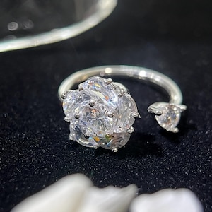 May include: A silver ring featuring a large, multi-faceted, clear gemstone cluster in a flower shape. A smaller, heart-shaped stone is set on the open end of the ring. The ring is set against a black background.