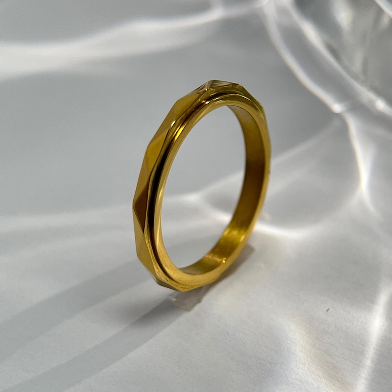 May include: A gold-colored ring with a geometric, faceted design. The ring has a band with multiple flat surfaces, creating a textured appearance. The ring is made of a shiny, metallic material.