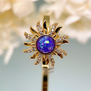 Könnte beinhalten: Ein goldfarbener Ring mit einem Sonnenstrahlen-Design. Der Ring hat einen zentralen, irisierenden blauen Edelstein, der von goldenen Strahlen und kleinen, klaren Edelsteinen umgeben ist. Der Ring ist vor einem weichen, verschwommenen Hintergrund platziert.