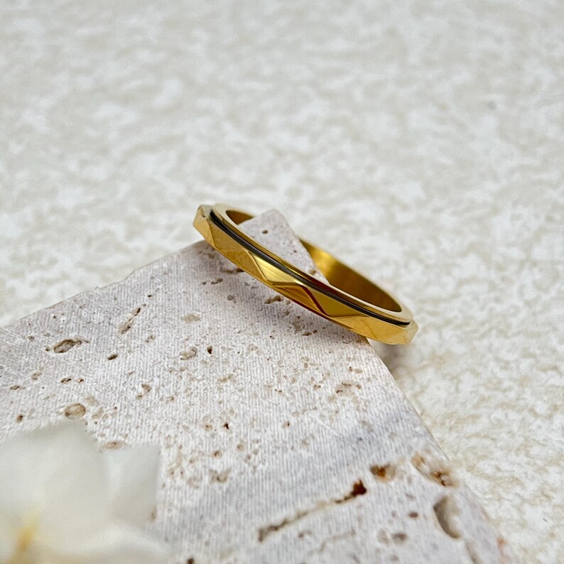 May include: A gold-colored ring with a geometric design. The ring has a series of angled facets that create a textured surface. The ring is resting on a light-colored, textured surface.
