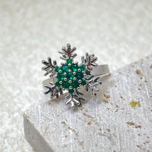 Könnte beinhalten: Ein silberner Ring mit einem Schneeflocken-Design. Die Mitte der Schneeflocke ist mit kleinen, runden, smaragdgrünen Steinen gefüllt, die von detaillierten, spitzen silbernen Zinken umgeben sind. Der Ring befindet sich vor einem neutralen, strukturierten Hintergrund.