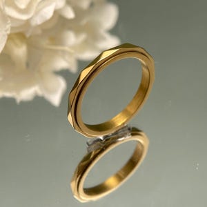 May include: A gold-colored ring with a geometric design. The ring has a faceted surface and a smooth inner band. The ring is reflected in a mirrored surface, with a blurred white flower in the background.