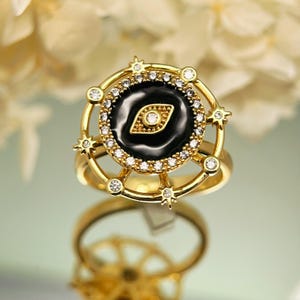 May include: A gold-colored ring with a black center featuring a gold-colored eye design. The ring is surrounded by small, clear gemstones and star-shaped accents. The ring is on a reflective surface.