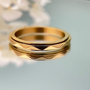 May include: A gold-colored ring with a geometric design. The ring has a textured, faceted band and a smooth, rotating center band. The ring is displayed on a reflective surface, with a soft-focus background.