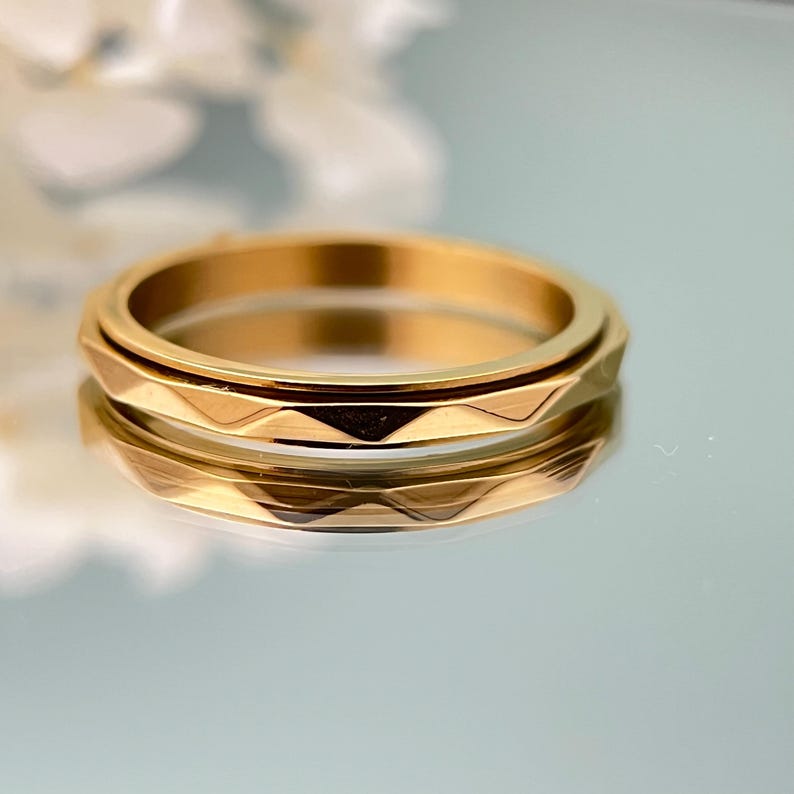 May include: A gold-colored ring with a geometric design. The ring has a textured, faceted band and a smooth, rotating center band. The ring is displayed on a reflective surface, with a soft-focus background.