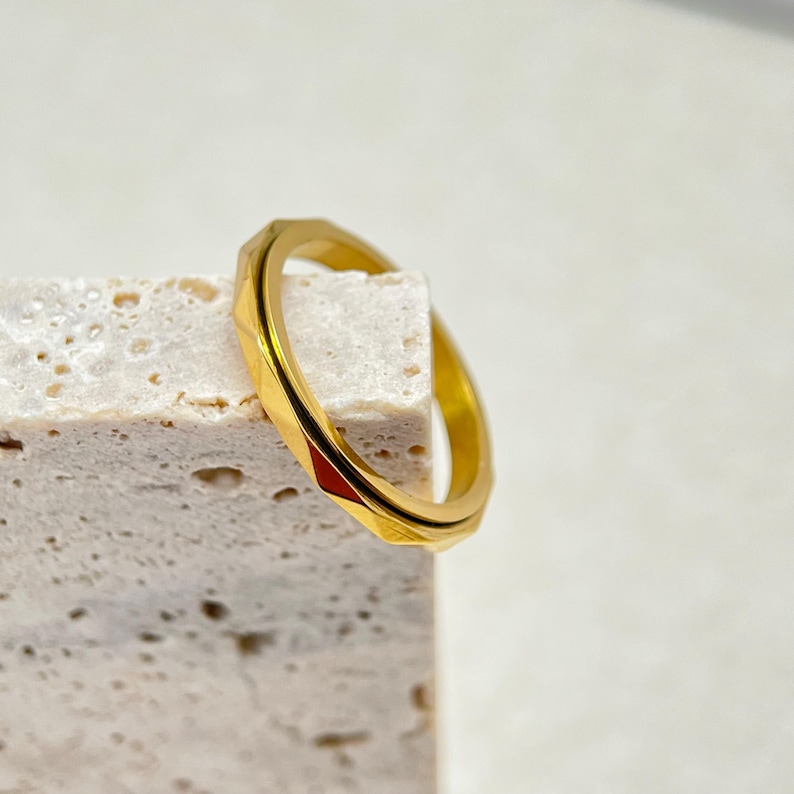 May include: A gold-colored ring with a geometric, faceted design. The ring is displayed on a textured, light-colored surface. The ring's design features multiple angled facets, creating a play of light and shadow.
