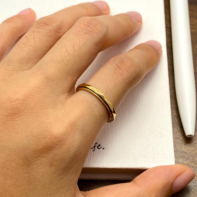 May include: A gold-colored ring with a geometric design is worn on a finger. The ring has a faceted surface, reflecting light. The ring is on a finger, with a notebook and pen in the background.