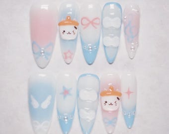 May include: A set of ten long, almond-shaped artificial nails. Each nail features a pastel color scheme of pink and blue, with various designs including bows, stars, clouds, and a cat-shaped character. Some nails have small, silver embellishments.