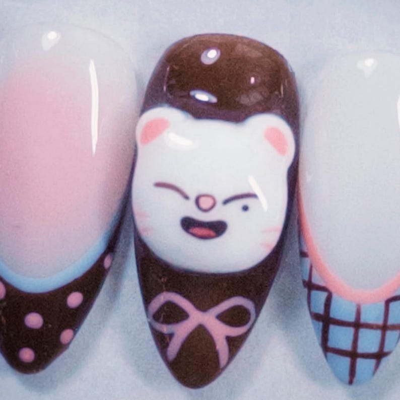 May include: Close-up of three nail art designs. One nail features a smiling white cat face on a brown background with a pink bow. The other nails have pink and blue designs. The nails are oval-shaped and have a glossy finish.