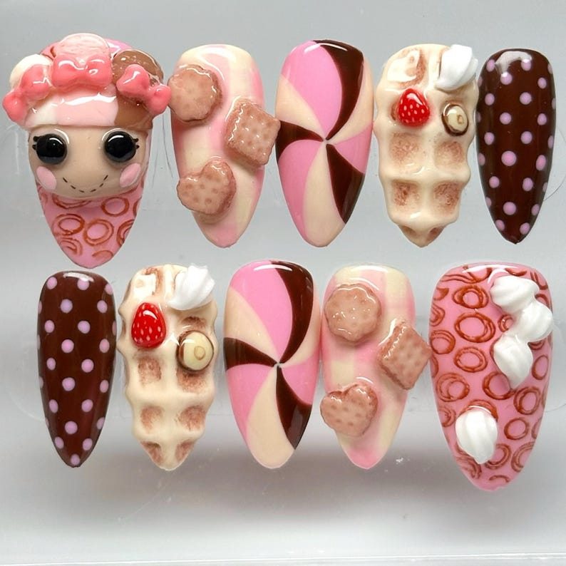 May include: A set of ten artificial nails with various designs. The designs include a pink sheep, cookies, hearts, pinwheels, waffles with strawberries, and polka dots. The color palette is primarily pink, brown, and white.