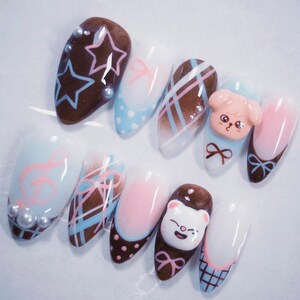 May include: A set of ten almond-shaped artificial nails with various designs. The nails feature a range of colors, including brown, pink, blue, and white, with designs such as stars, bows, and cartoon characters. Some nails have pearl accents.