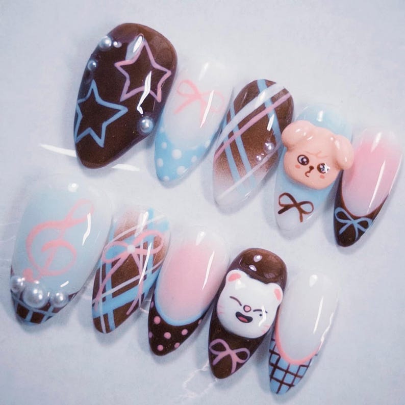 May include: A set of ten almond-shaped artificial nails with various designs. The nails feature a range of colors, including brown, pink, blue, and white, with designs such as stars, bows, and cartoon characters. Some nails have pearl accents.