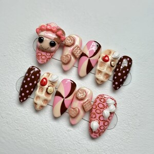 May include: A set of ten almond-shaped press-on nails with a dessert theme. Designs include pink and brown swirls, waffle and cookie accents, and a nail with a pink face and a hat. Some nails have a brown polka dot pattern.